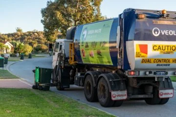 A waste collection truck with a green waste bin being lifted by a mechanical arm in a suburban neighborhood during daylight.